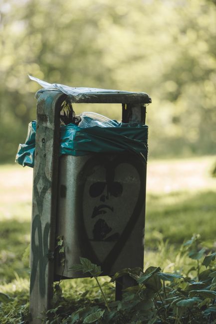 A weathered metal waste bin situated in a garden or park area, featuring a graffiti-style image of a face with a sad expression painted on its front. The bin has a slightly rusted surface with patches