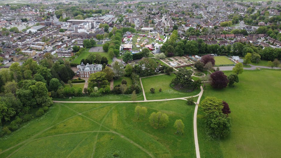 A garden pathway in Raphael Park, Gidea Park, lined with vibrant flowering plants and lush green foliage on either side. Overhead, a black metal arbor arch is covered with climbing plants, creating a 