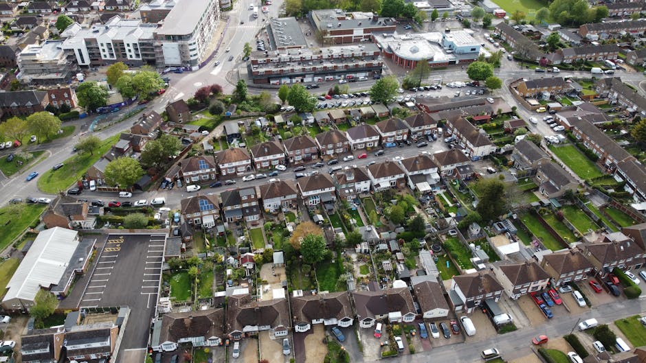 An aerial view of a residential neighborhood showing rows of semi-detached houses with small, well-maintained gardens featuring lush green lawns, neatly trimmed hedges, and a variety of trees with ful